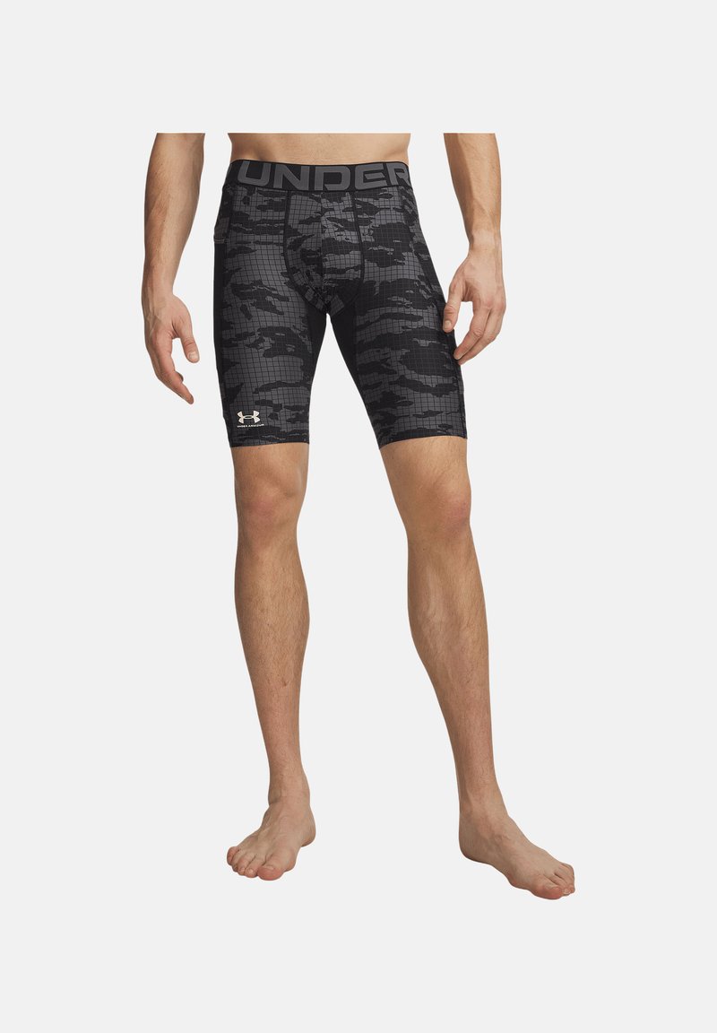 Homme portant un short de compression camouflage noir et gris avec un large texte sur la taille et le logo blanc Under Armour sur la cuisse gauche.