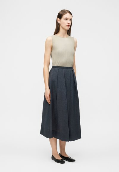 Γυναίκα που στέκεται φορώντας γιλέκο μπεζ χωρίς μανίκια με ριμπ ύφασμα και μακριά midi φούστα σε σκούρο μπλε με ψηλή μέση, φορώντας μαύρα φλατ παπούτσια, πάνω σε απλό φόντο.