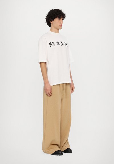 White t-shirt with black block lettering, paired with wide beige pants and black shoes. Textured fabric and relaxed fit design.