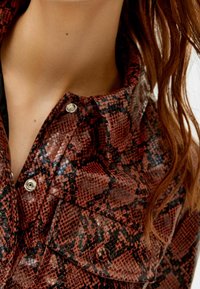 Chemise en imprimé serpent rouge et noir, avec une finition texturée, un col et des boutons-pression en argent. La vue en gros plan met en évidence le détail du tissu.