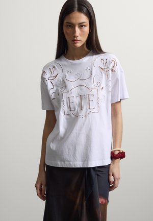 Junge Frau mit langen Haaren, die ein weißes, ausgeschnittenes besticktes T-Shirt mit dem Schriftzug „ETE“ und einen dunklen Rock trägt, posiert vor einem schlichten Hintergrund.