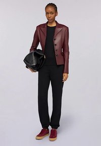 Giacca in ecopelle bordeaux con taglio sartoriale, maglione nero sotto, pantaloni neri e sneakers color marrone scuro. Borsa nera in mano.