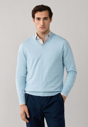 Joven con un suéter de cuello en V azul claro sobre una camisa de rayas con cuello, de pie con una mano en el bolsillo de sus pantalones azul marino.