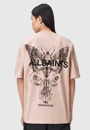 Osoba w bladoróżowej, oversize'owej koszulce z czarną grafiką szczegółowego motyla i czaszki zwierzęcia oraz czarnym napisem "ALLSAINTS LONDON THE SAINTHOOD" na plecach.
