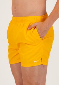 Geltonos Nike maudymosi šortai su elastinga juosta, šoninėmis kišenėmis ir baltu logotipu. Lygi tekstūra, ilgio iki kelių su įpjovomis.