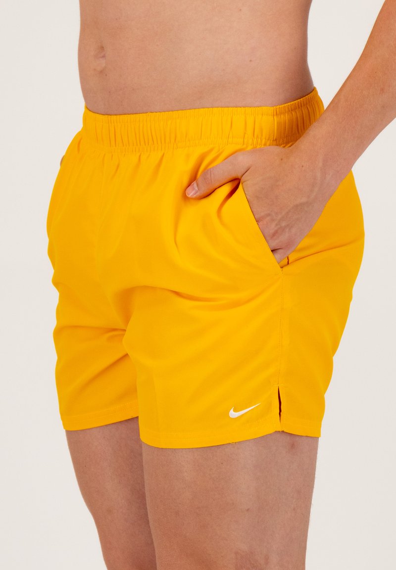 Żółte spodenki pływackie Nike z elastycznym paskiem, bocznymi kieszeniami i białym logo. Gładka tekstura, długość powyżej kolana z rozcięciami.