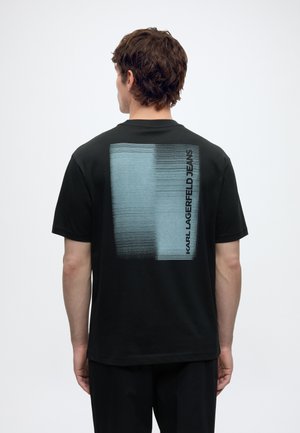 Muškarac smeđe kose u crnoj majici pokazuje veliki plavo-crni pikselirani kvadrat s tekstom "KARL LAGERFELD JEANS" na leđima.