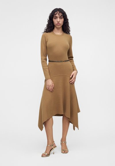 Un mannequin se tient devant un fond blanc uni, vêtu d’une robe en maille brun camel à ourlet asymétrique, avec une ceinture chaîne et des talons dorés.
