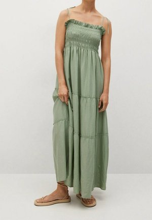 Femme portant une robe maxi à volants verte clair sans manches, avec un corsage smocké et de fines bretelles, associée à des sandales plates beiges.