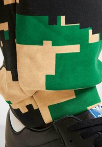 Camuflerede sokker i grøn, sort og beige stof, parret med en sort sneaker med et hvidt logo og snørredetaljer.