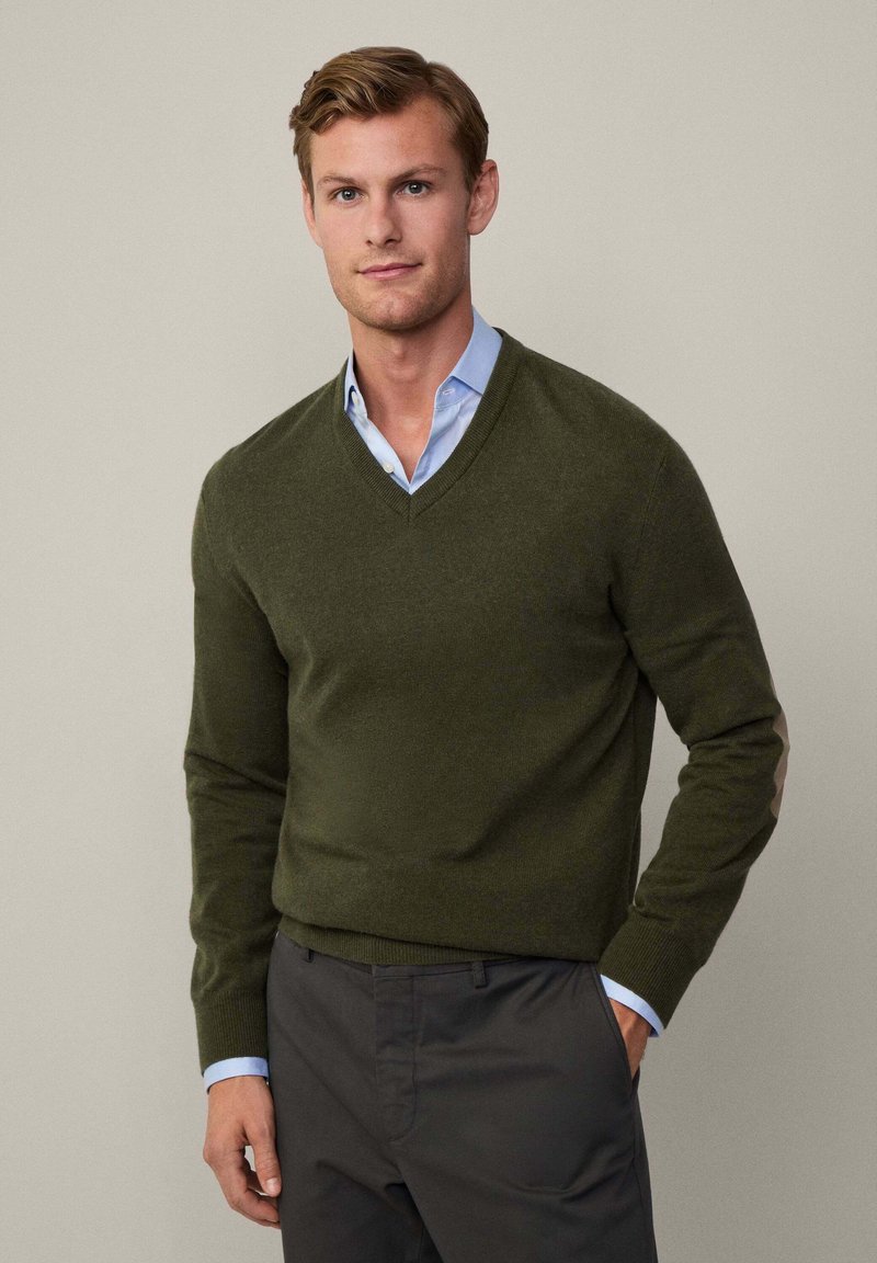 Olive green v-neck sweater in soft fabric, featuring elbow patches. Worn over a light blue collared shirt, paired with dark trousers.