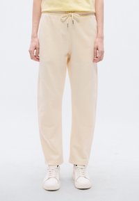 Παντελόνια φόρμας σε κρεμ απόχρωση με κορδόνι στη μέση, τσέπες στο πλάι και άνετη γραμμή, συνδυασμένα με λευκά αθλητικά παπούτσια. Ομαλή υφή και ίσια μπατζάκια.