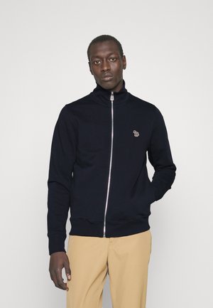 Sweat-shirt zippé bleu marine avec col haut, poignes côtelées, logo brodé sur la poitrine et tissu lisse. Assorti à un pantalon beige.