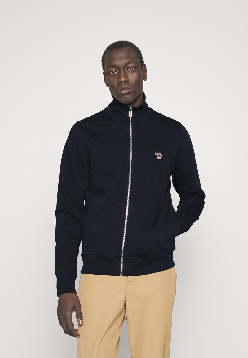 Sweat-shirt zippé bleu marine avec col haut, poignes côtelées, logo brodé sur la poitrine et tissu lisse. Assorti à un pantalon beige.