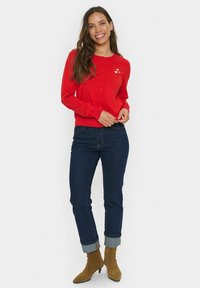 Cardigan rouge avec des boutons, orné d'une broderie en forme de cœur. Associe à un jean sombre et des bottines à lacets beiges. Texture lisse et design ajusté.