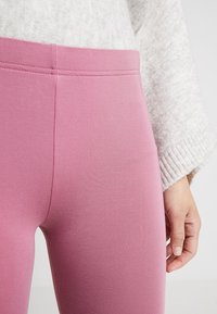 Närbild av en person som bär högmidjade rosa leggings och en vit stickad tröja, med höger hand avslappnad vid sidan.