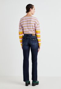 Striped purple and yellow long-sleeve top, dark blue denim flared jeans, and green chunky heeled shoes; Lee logo visible on waistband.