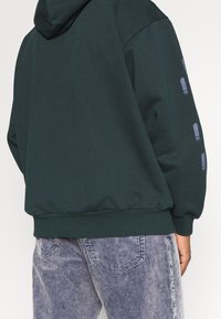 Tmavě zelená mikina s kapucí a volným střihem, s texturovanými akcenty na rukávech a žebrovanými manžetami. Doplněná pruhovanými kalhotami.