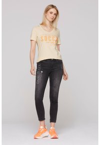 Camiseta de cuello en V beige con texto gráfico naranja, combinada con jeans ajustados negros que tienen acentos impresos en blanco. Modelo lleva zapatillas naranjas.