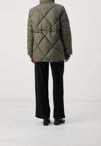 Oliwkowa pikowana kurtka puchowa z przeszyciami w kształcie diamentów, wysokim kołnierzem i elastycznymi mankietami, zestawiona z czarnymi spodniami o szerokich nogawkach i grubymi butami na obcasie.