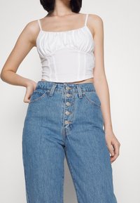 Haut blanc, court, à volants avec des fines bretelles, associé à un jean taille haute, bleu clair, comportant une fermeture à cinq boutons et des poches avant.