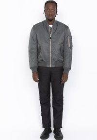 Giubbotto bomber grigio con polsini e vita a coste, chiusura lampo frontale, tasche laterali e brandizzazione discreta; abbinato a pantaloni neri e stivali.