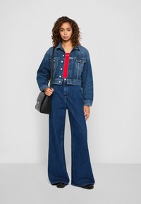 Kvinne som står mot en ensfarget bakgrunn, iført en kort denimjakke, vide jeans, rød topp, svarte sko, og bærer en svart skulderveske.