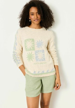 Frau mit lockigem Haar, die einen cremefarbenen Pullover mit Sonnen- und Wellenmustern und hellgrüne Shorts trägt, stehend vor einem weißen Hintergrund.