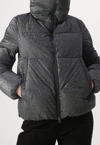 Svart puffjacka med en glänsande texturfinish, dragkedjestängning och två knäppta fickor. Har också en huva och stygnpanel design.