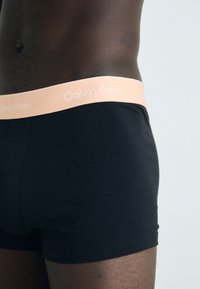 Crne bokserice s glatkom teksturom i elastičnom etiketom koja sadrži svijetlo ružičasti "Calvin Klein" logo. Jednostavan, uski dizajn.