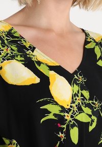 Tela negra con un vibrante patrón de limones y hojas verdes. Diseño de cuello en V con una textura suave. Vista cercana de los detalles de la prenda.