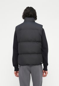 Homem com cabelo escuro de comprimento médio, vestindo um colete acolchoado preto sobre uma camiseta de manga longa preta e calças cinzas, de pé de costas.