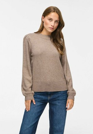 Femme aux longs cheveux bruns portant un pull en tricot beige avec des manches bouffantes et un jean bleu sur un fond blanc uni.