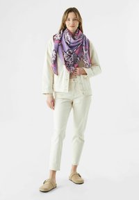 Foulard triangle surdimensionné en violet et rose avec des accents de léopard. Le modèle porte une veste en denim blanche et un pantalon crème avec des chaussures beiges.