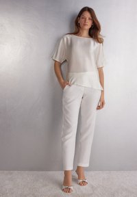 Blusa de seda blanca con un escote ancho y mangas cortas, combinada con pantalones blancos a medida. La modelo lleva zapatos de punta abierta de color claro.