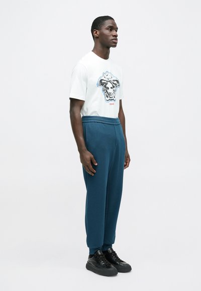 Biele tričko s grafickým dizajnom hlavy Medúzy; v kombinácii s tyrkysovými teplákmi a čiernymi teniskami. Jednoduchý okrúhly výstrih a voľný strih.