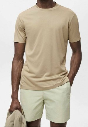Mann steht vor einem schlichten Hintergrund, trägt ein beiges T-Shirt und helle Khaki-Shorts und hält eine zusammengefaltete Jacke in der linken Hand.