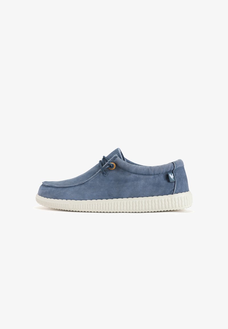 Blue canvas slip-on shoe with a round toe, white rubber outsole, and contrasting decorative stitching. Features a small logo tag.