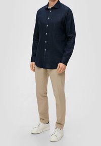 Camicia a maniche lunghe blu navy in tessuto leggero, con colletto button-down, bottoni frontali e vestibilità relaxed, abbinata a pantaloni beige e scarpe bianche.