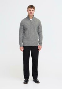 Grijze zip-up trui gemaakt van gebreide stof, gecombineerd met zwarte broeken en zwarte schoenen, staand voor een effen achtergrond.