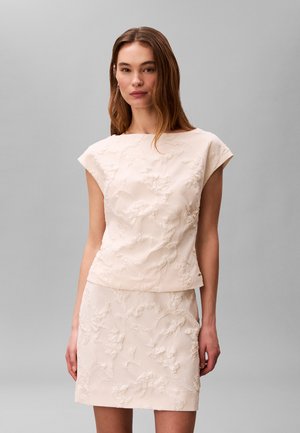 Kobieta w bezrękawnikowej, kremowej, teksturowanej sukience z kwiatowym wzorem, stojąca na jednolitym szarym tle.