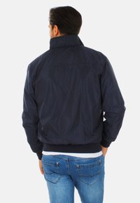 Giacca bomber blu navy con colletto alto, polsini elasticizzati e una trama liscia. Indossata su jeans di denim azzurro.