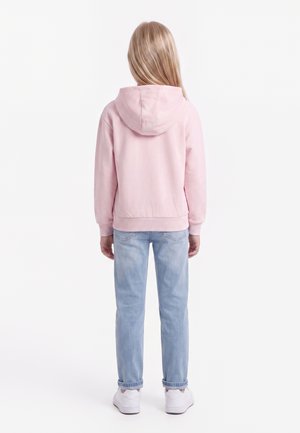 Enfant aux longs cheveux blonds portant un sweat à capuche rose, un jean bleu clair et des baskets blanches, debout de dos sur un fond blanc.