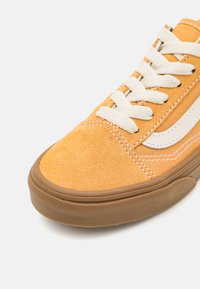 Zapatilla de ante amarilla con cordones y costuras blancas, que cuenta con una suela de goma y un logo lateral en blanco. Punta redondeada y parte superior texturizada.