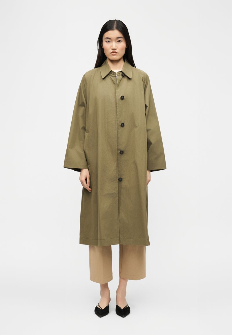 Γυναίκα με μακριά σκούρα μαλλιά που φοράει ένα παλτό χακί με κουμπιά πάνω από μπεζ κοντά παντελόνια και μαύρα παπούτσια με μυτερή μύτη, στέκεται μπροστά σε ένα απλό φόντο.