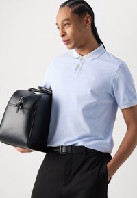 Svart lærveske med rund form og glidelås, kombinert med en lyseblå polo skjorte og svarte bukser; minimal logo på skjorten.