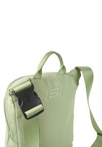 Mochila de nylon verde con correa ajustable, asa superior y hebilla de plástico negra. Presenta un logo grabado en la parte delantera.