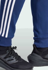 Zwarte sportschoenen met een geprofileerde mesh bovenkant en rubberen zolen, gecombineerd met marineblauwe trainingsbroek met drie witte strepen aan de zijkant.