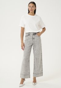 Hvid kortærmet top med nagledetaljer, parret med højtaljede, vide grå denimjeans og sølvfarvede spidse hæle.