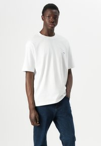 White cotton T-shirt with a crew neck, short sleeves, and a blue printed design on the left chest. Accompanied by dark blue denim jeans.